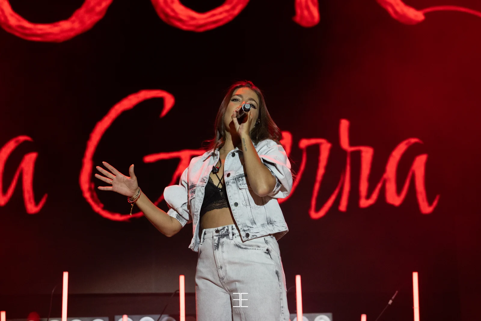Ana Guerra canta con un brazo extendido hacia el público, con el neón rojo detrás