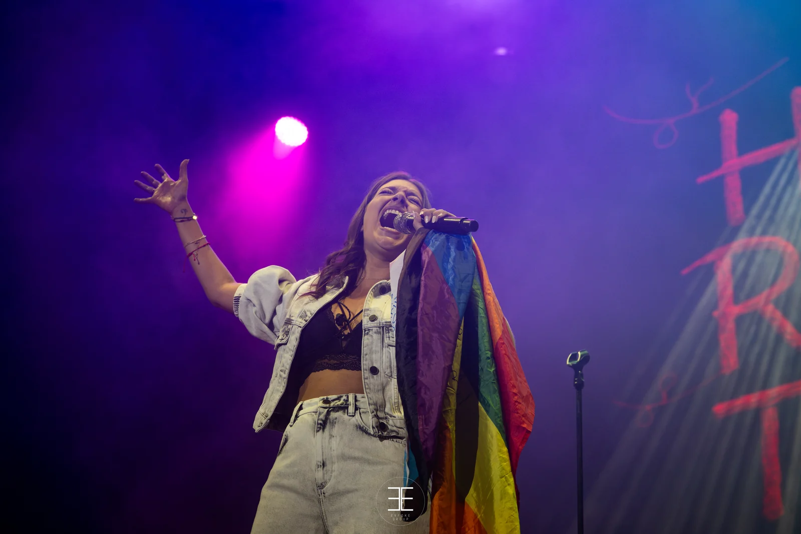 Ana Guerra canta con una bandera arcoíris sobre el hombro y el brazo abierto, bajo una luz morada