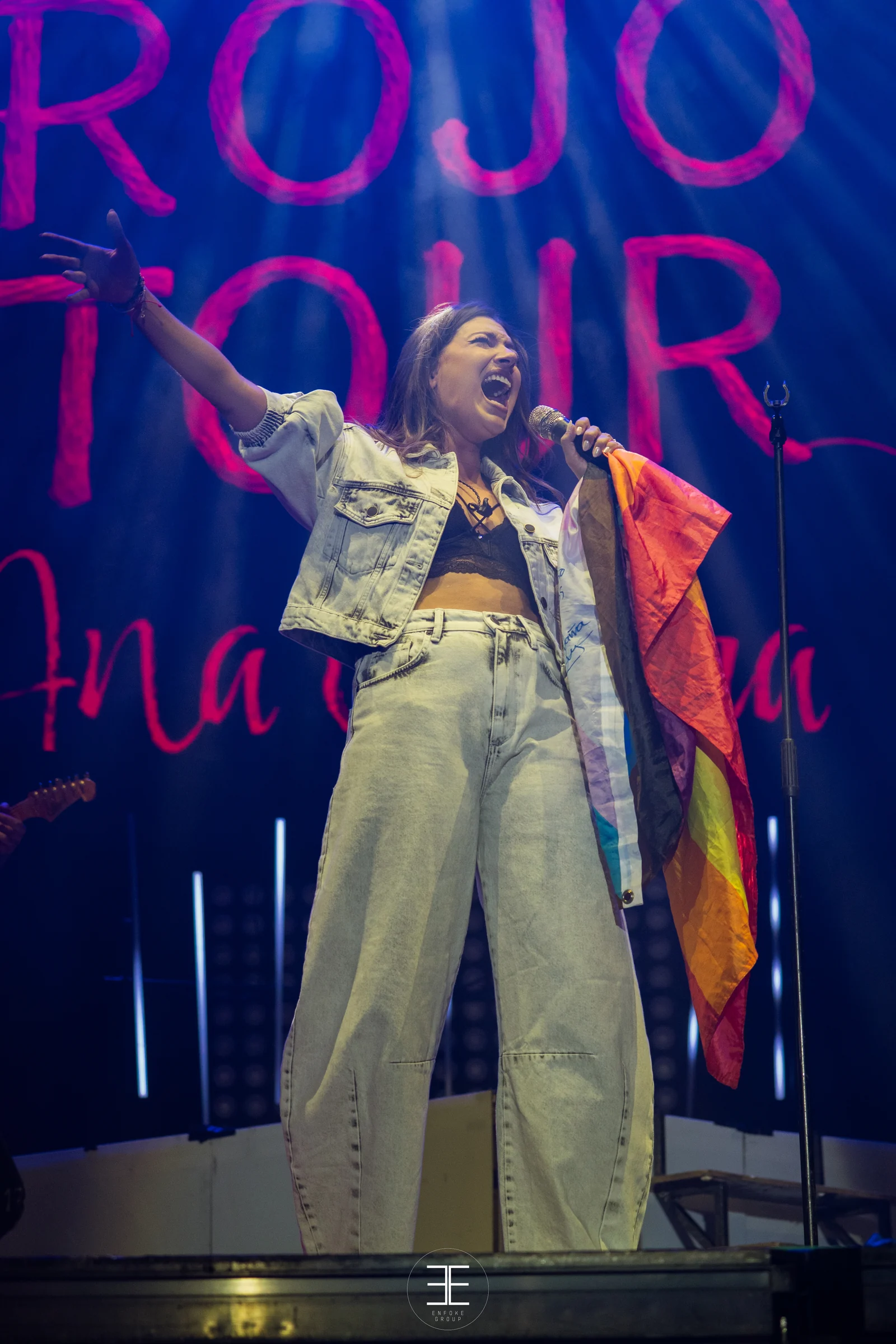 Ana Guerra, de cuerpo entero y en vertical, canta con la bandera arcoíris ante el rótulo de neón de su gira