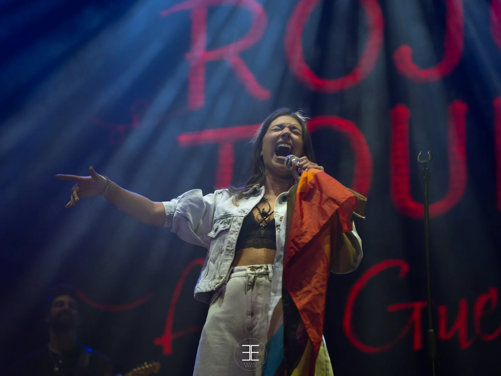 Ana Guerra canta señalando al frente con la bandera arcoíris sobre el hombro y el neón rojo detrás