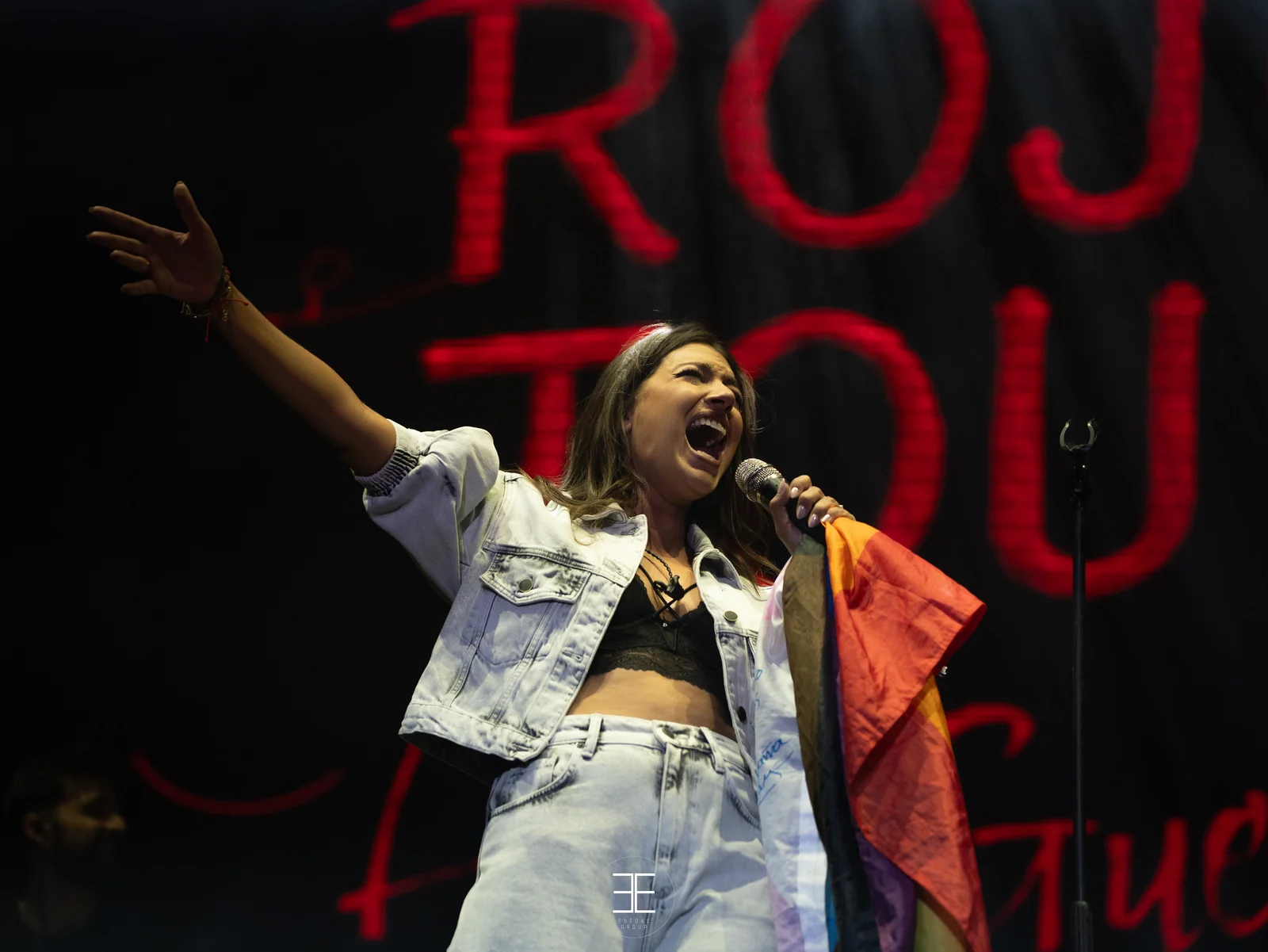 Ana Guerra levanta el brazo cantando con la boca muy abierta, con la bandera arcoíris al hombro