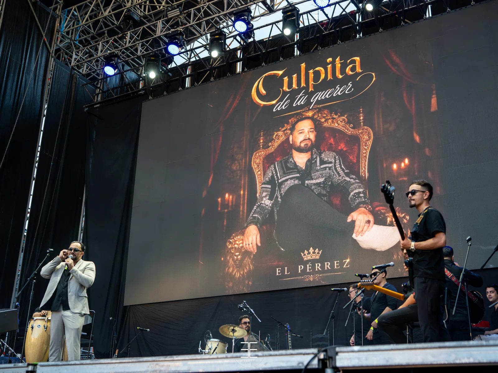 Plano general del escenario con la portada del disco en la pantalla gigante; El Pérrez canta abajo a la izquierda junto a una conga