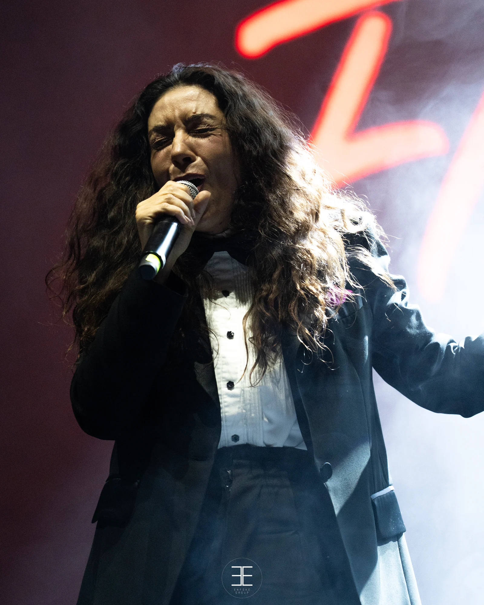 Sandra Carrasco canta al micro con el brazo derecho abierto hacia fuera del cuadro, melena rizada sobre la cara y el rótulo rojo desenfocado detrás