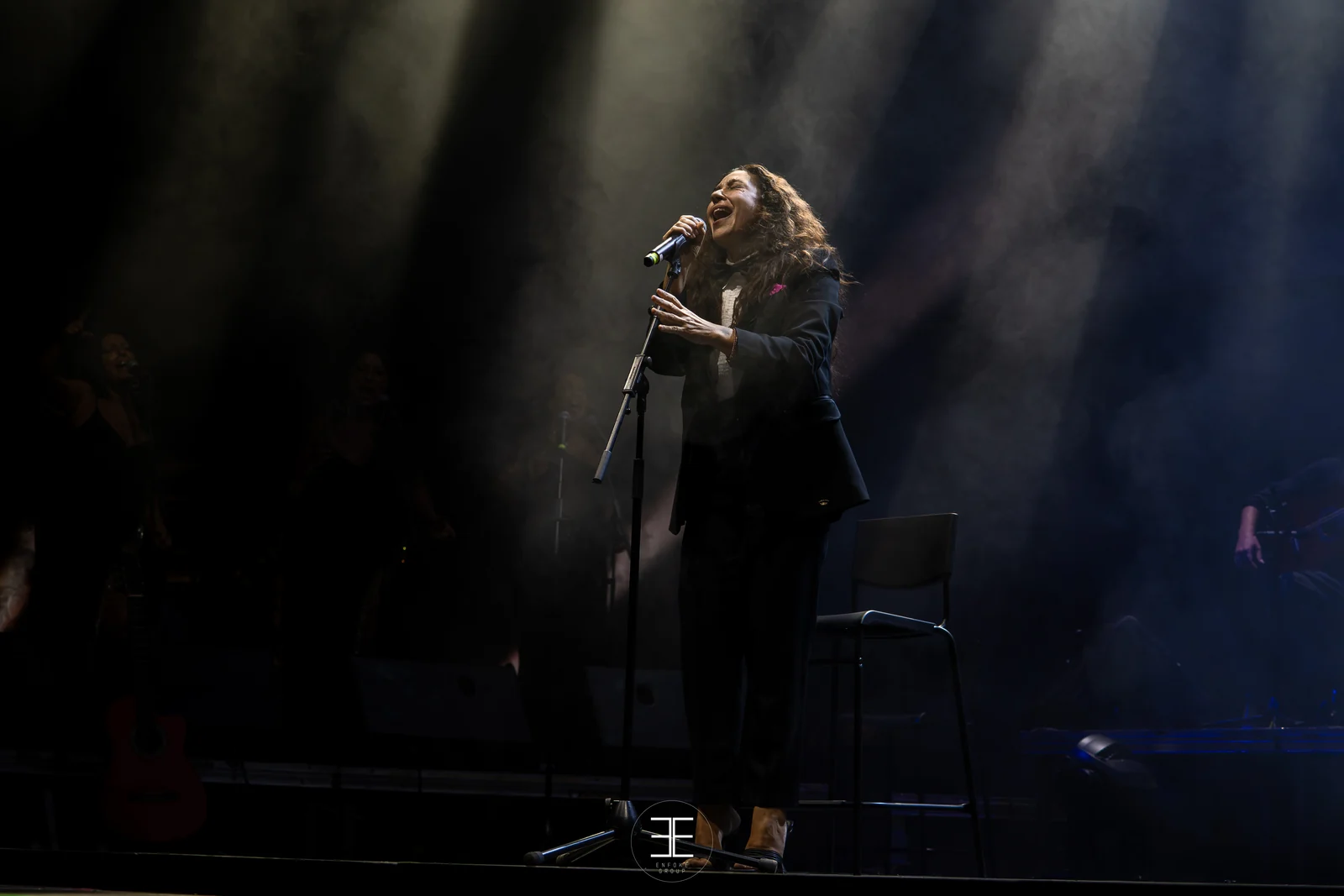 Plano general del escenario: Sandra Carrasco ante el micrófono con la cabeza echada atrás y el resto del grupo en penumbra