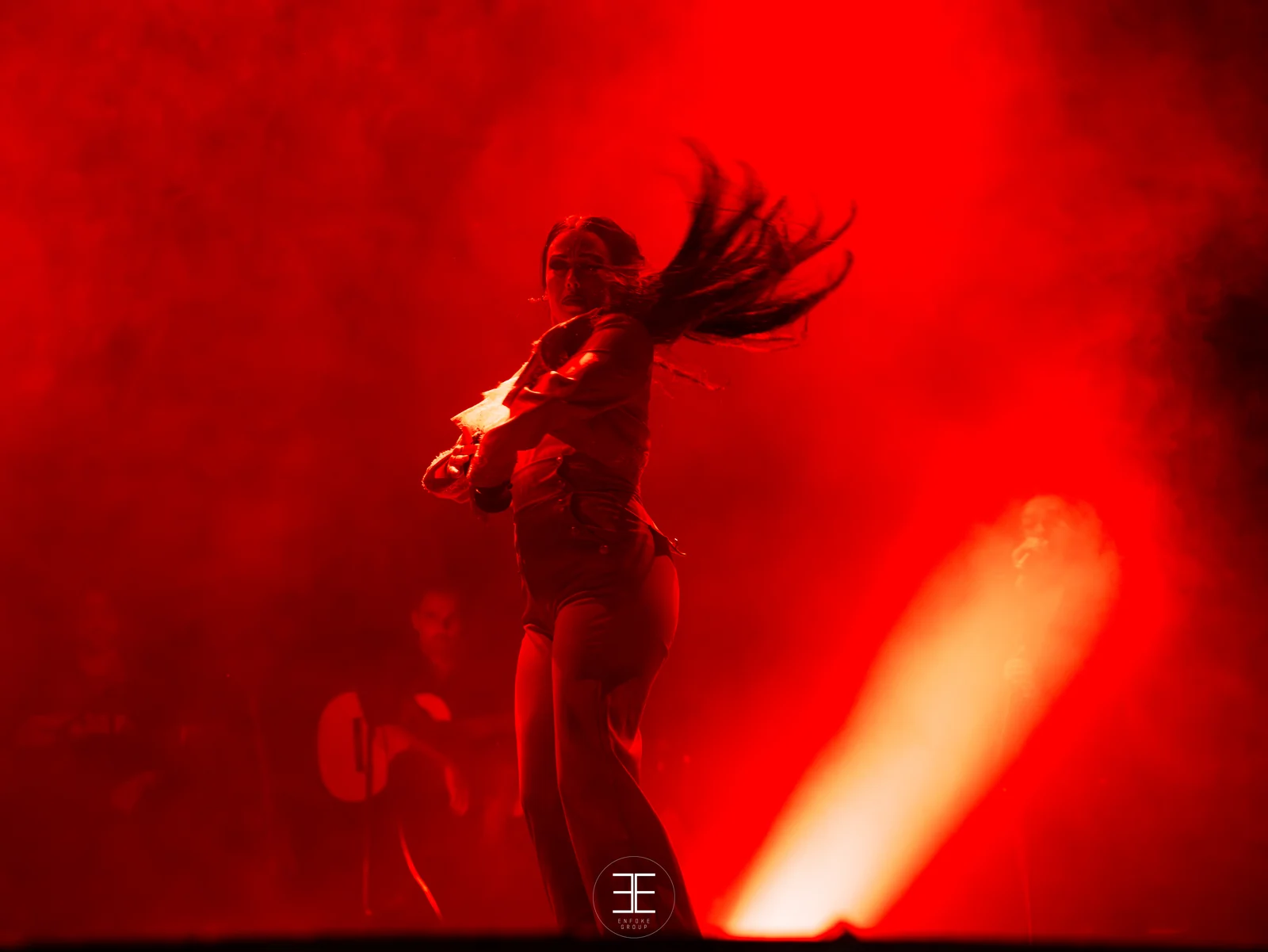 Belén López girando a contraluz rojo con la melena suelta en el aire; al fondo, un guitarrista sentado y un cono de luz