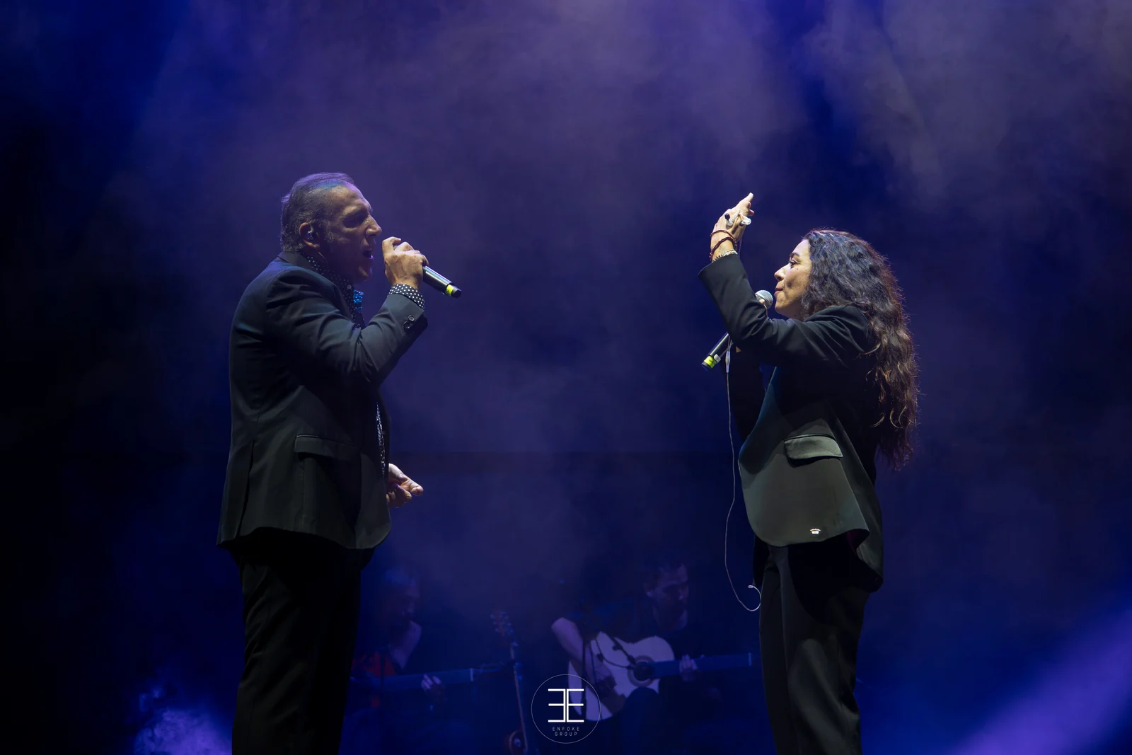 Enrique Heredia «Negri» y Sandra Carrasco, frente a frente con sus micrófonos bajo luz azul; detrás, dos guitarristas sentados