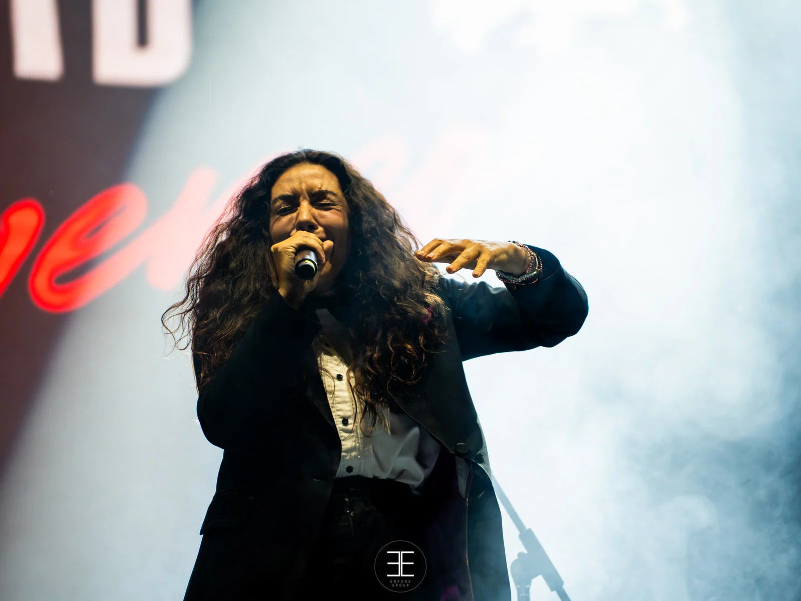 Sandra Carrasco canta con micro de mano y la mano derecha levantada; americana negra sobre camisa blanca y el rótulo rojo del espectáculo al fondo