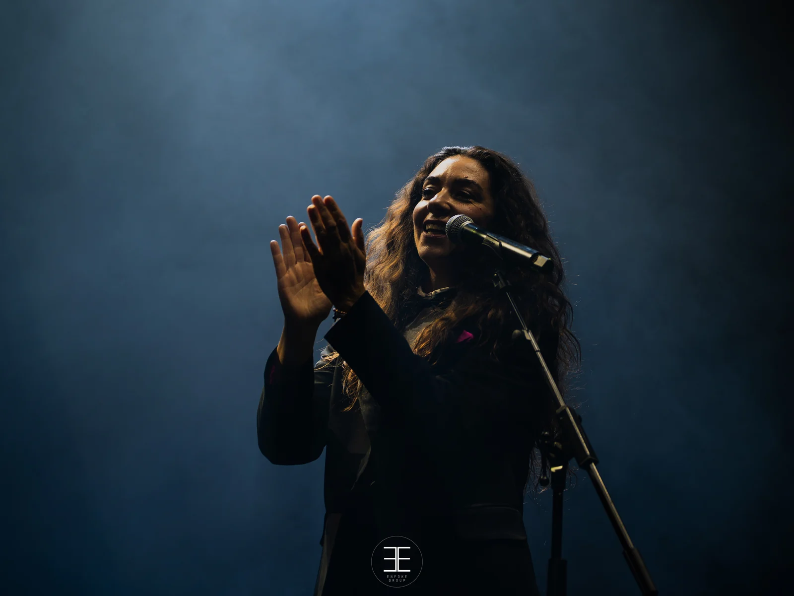 Sandra Carrasco junta las palmas a la altura del pecho, al lado del micro de pie, sobre fondo azul de humo