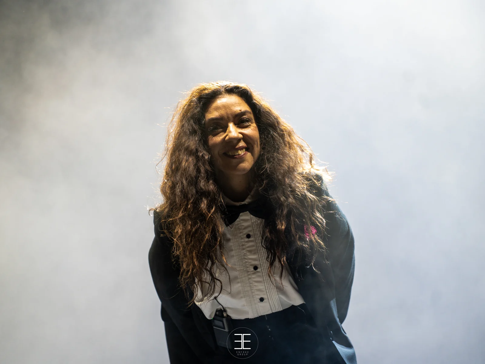 Retrato de Sandra Carrasco sonriendo, con las manos a la espalda, recortada sobre una pared de humo blanco iluminado