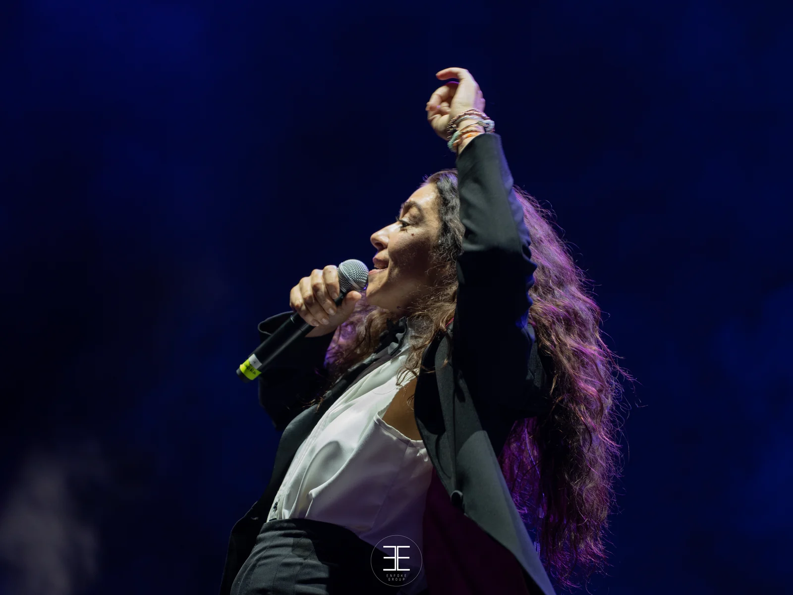Sandra Carrasco con el brazo derecho alzado y el puño cerrado mientras canta, pulseras en la muñeca y camisa blanca bajo la americana