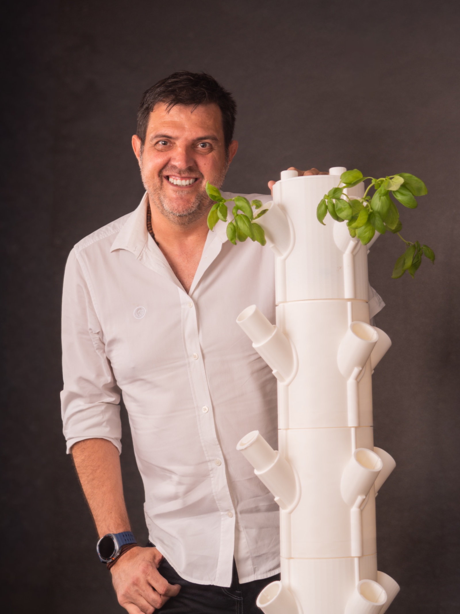 Fernando Fernández Martínez sonriendo con camisa blanca y la mano sobre una torre de cultivo blanca con plantas, sobre fondo oscuro