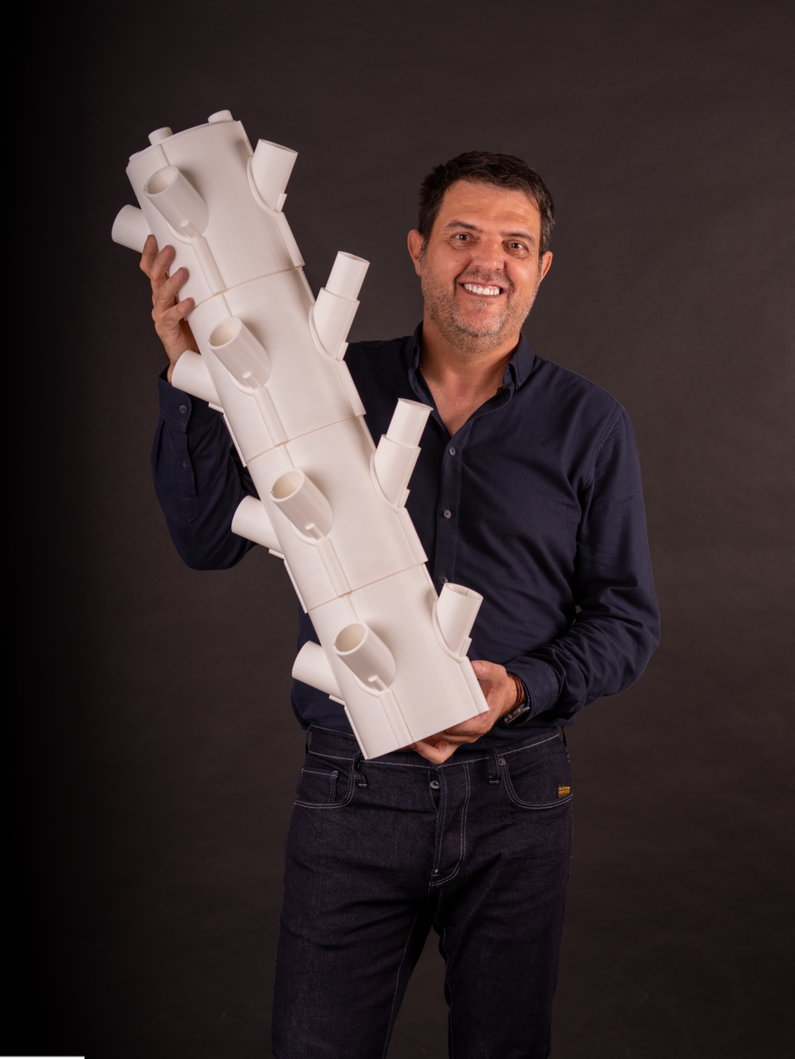 Fernando Fernández Martínez con camisa azul marino sosteniendo en diagonal una torre de cultivo blanca, sobre fondo oscuro