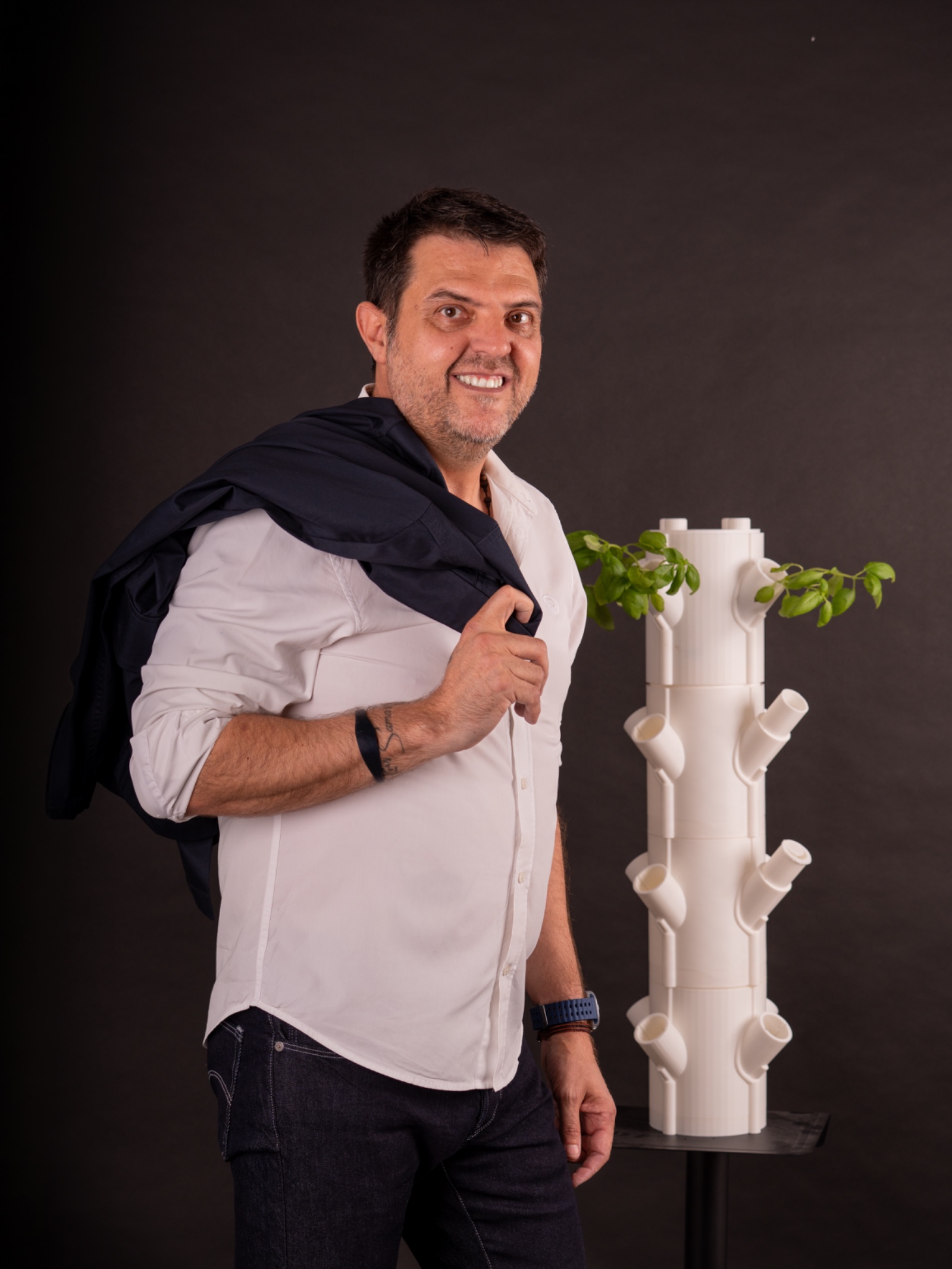 Fernando Fernández Martínez con camisa blanca y la americana al hombro, junto a una torre de cultivo con plantas