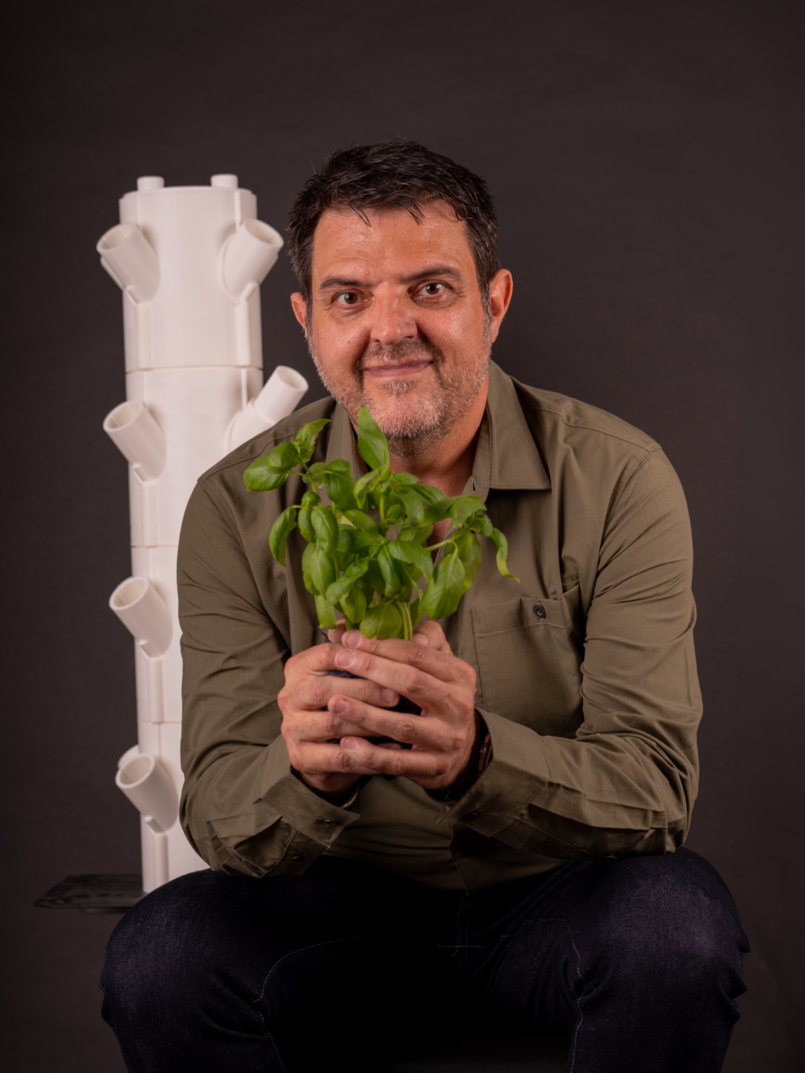 Fernando Fernández Martínez con camisa verde oliva sujetando una planta con las dos manos, con la torre de cultivo detrás