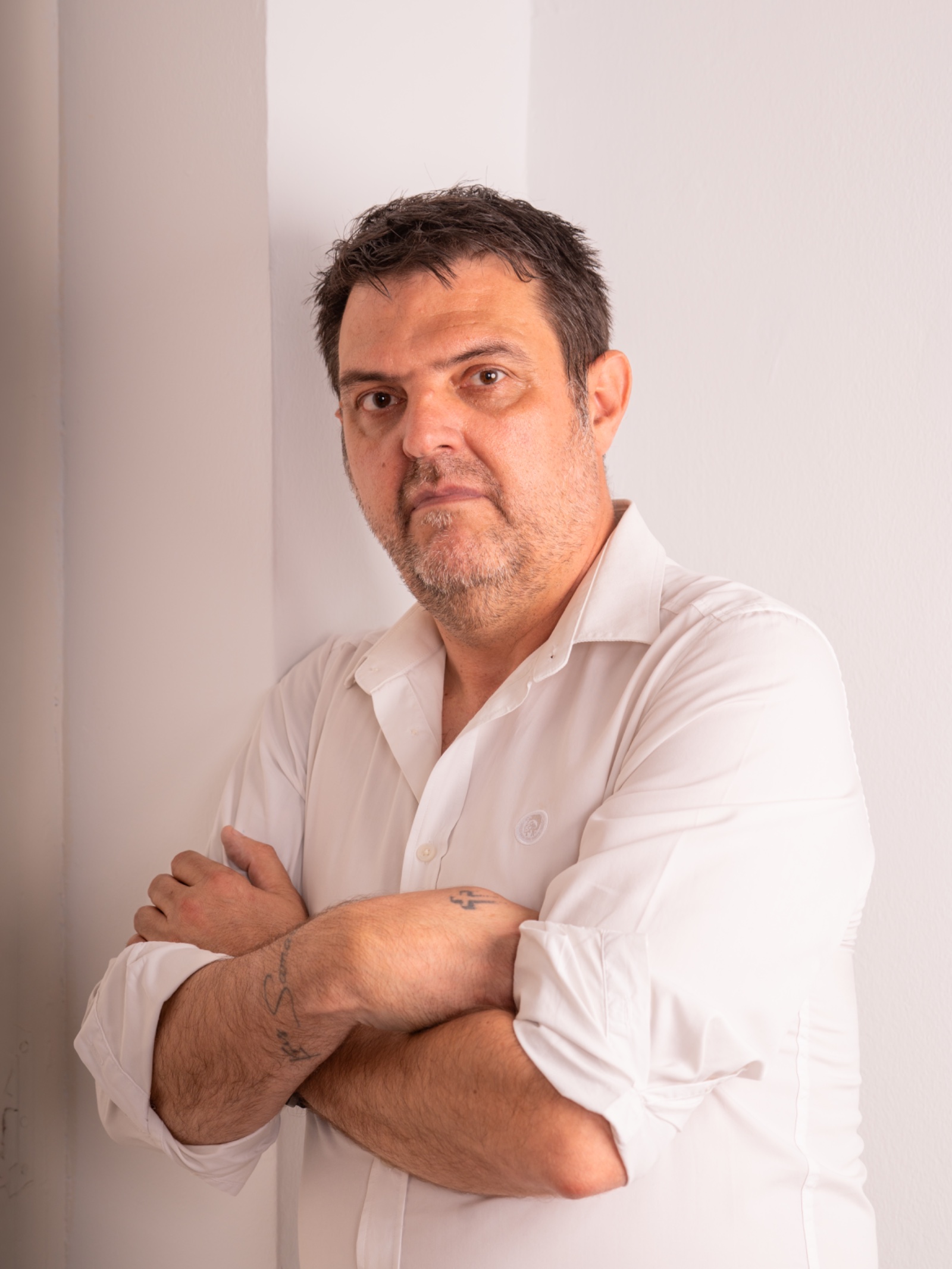 Fernando Fernández Martínez con camisa blanca y los brazos cruzados, apoyado en una pared clara y mirando a cámara