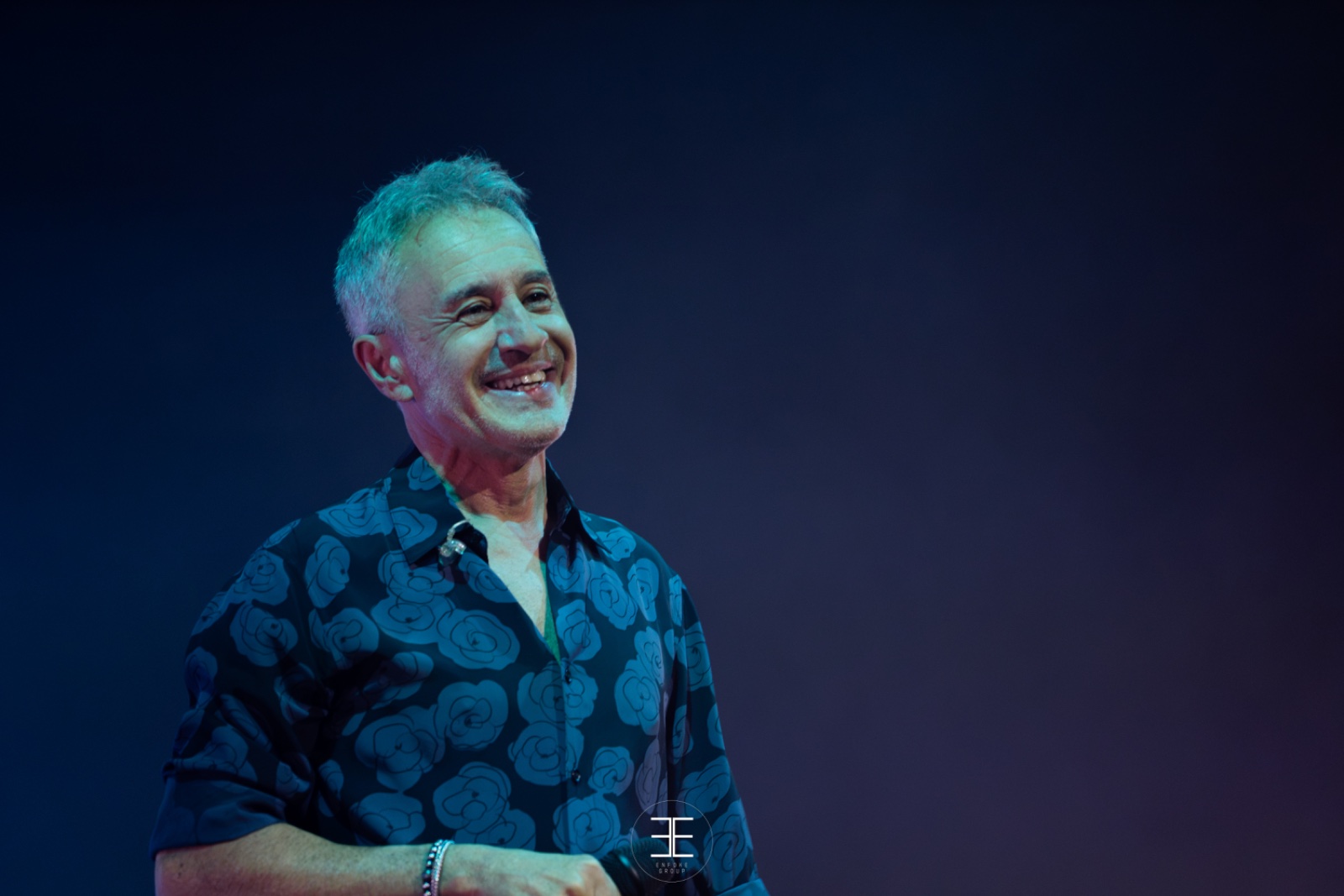 Sergio Dalma sonríe mirando a un lado, con camisa oscura estampada de flores, sobre fondo azul