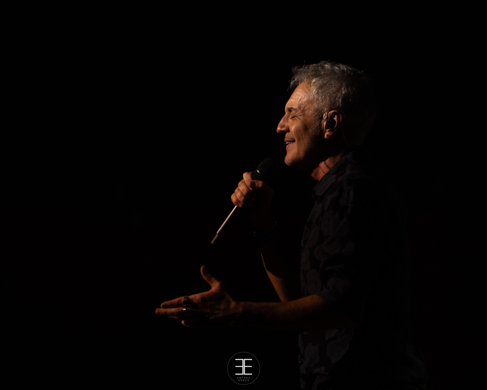 Sergio Dalma de perfil, sonriendo en la penumbra, con la mano abierta